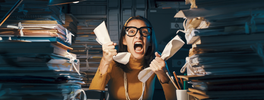 Angry stressed woman sitting at office desk screaming and crumpling paperwork, she is overloaded with work